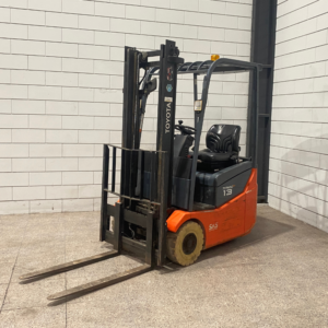 Een oranje elektrische heftruck van het merk Toyota, model 7FBE15, staat op een betonnen vloer in een magazijnomgeving. De heftruck heeft een eenvoudige constructie met een bestuurdersstoel en een vorkenframe.