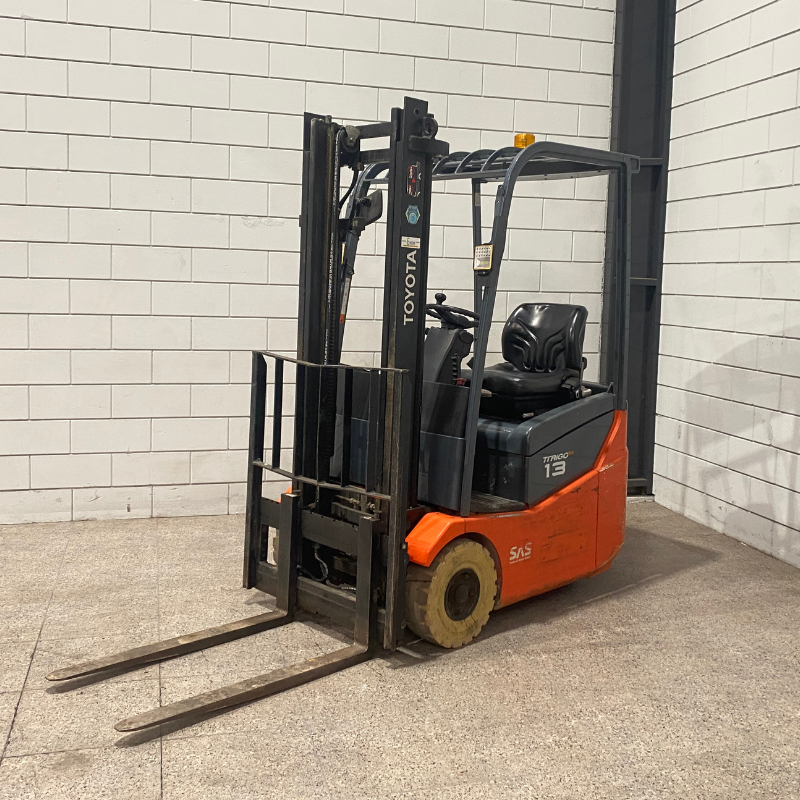 Een oranje elektrische heftruck van het merk Toyota, model 7FBE15, staat op een betonnen vloer in een magazijnomgeving. De heftruck heeft een eenvoudige constructie met een bestuurdersstoel en een vorkenframe.